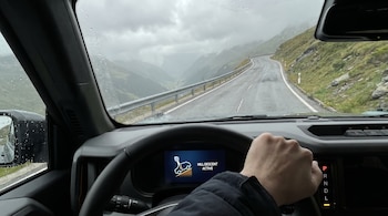 Vista del interior de un coche descendiendo una carretera de montaña. Pantalla muestra 'Hill Descent Active', palanca en modo 'L'. Mano en volante. Carretera mojada, cielo nublado.