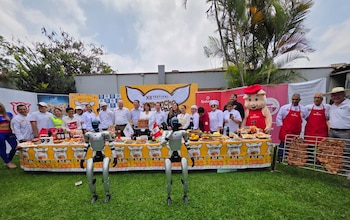 Un grupo de personas, cocineros, dos robots humanoides y una botarga posan detrás de mesas con comida y una parrilla en un jardín
