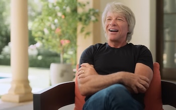 Jon Bon Jovi, un hombre de cabello gris con camiseta negra y vaqueros, sentado con los brazos cruzados en un sillón, sonriendo y riendo