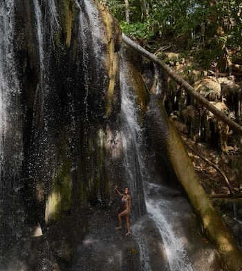 Una mujer en traje de baño de dos piezas permanece bajo una cascada en medio de rocas y vegetación tropical