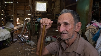 Un hombre mayor apoya su mano sobre un bastón dentro de una habitación con paredes de madera, suelo de tierra y enseres domésticos.