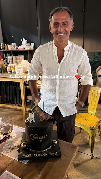 Hombre sonriente, camisa blanca, pastel negro con velas, figuras de dos perros, copa de vino, mesa de madera. Textos: Feliz Cumple Papá, Feliz cumple @dflatorre
