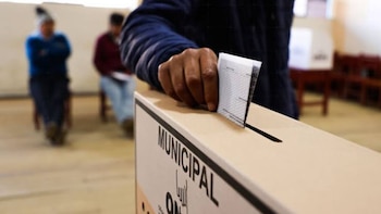 Una mano introduce una cédula de votación doblada en una urna de cartón con el logotipo de la ONPE