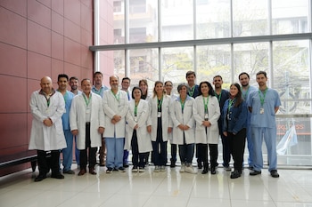 Un grupo de dieciséis personas con batas blancas y uniformes médicos de color azul posan alineadas en un interior frente a un gran ventanal