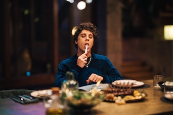 Hombre joven con cabello rizado sentado en una mesa, sosteniendo un objeto blanco con la boca, rodeado de platos, ensaladera y vasos en ambiente oscuro