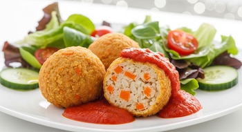 Primer plano de tres albóndigas doradas de zanahoria y arroz en plato blanco. Una está partida, mostrando el interior. Llevan salsa de tomate y ensalada.