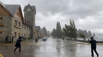 Personas con abrigo y paraguas caminan por una calle mojada frente a edificios de piedra con torre de reloj bajo un cielo nublado y lluvia.