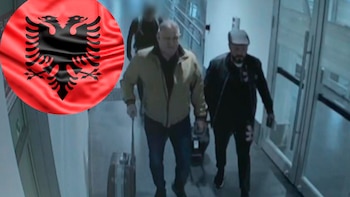 Bandera de Albania, circular y roja con águila bicéfala negra, superpuesta sobre tres hombres que caminan por un pasillo. Un hombre usa un bastón