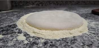 Primer plano de una masa redonda y plana, cubierta con harina, reposando sobre una encimera de granito oscuro salpicada de harina en polvo