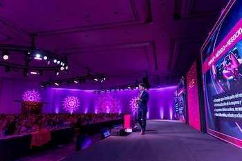 Un hombre en traje habla en un escenario morado con grandes pantallas y focos de techo, frente a una audiencia numerosa sentada en mesas