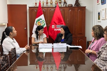 Cinco mujeres sentadas alrededor de una mesa de madera brillante. Dos de ellas, Melina Locatelli Alfaro y Deborah Revilla, se miran frente a banderas peruanas