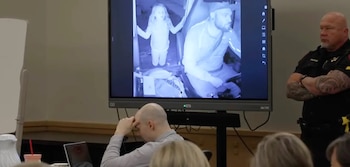 Una pantalla grande en una sala de audiencias muestra una imagen monocromática de una niña y un hombre barbudo en un vehículo, junto a un oficial y un hombre calvo