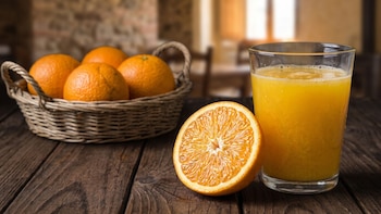 Un vaso de zumo de naranja, media naranja cortada y una cesta de mimbre con naranjas enteras sobre una mesa de madera