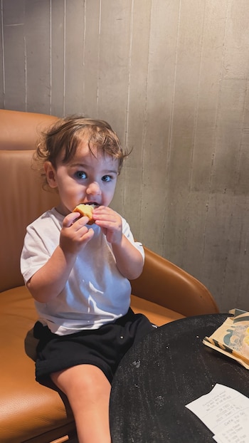 Niño sentado en silla de cuero marrón comiendo un trozo de alimento. Lleva camiseta blanca y pantalones negros. Sobre mesa negra hay una bolsa de papel y un recibo