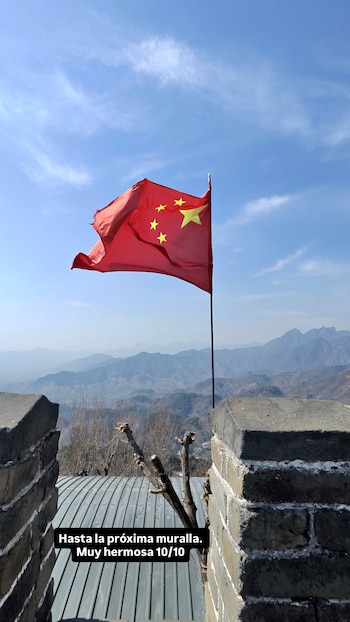 Bandera roja de China con cinco estrellas amarillas ondeando al viento sobre un fondo de montañas brumosas y cielo azul. Se ven partes de un muro de piedra en primer plano