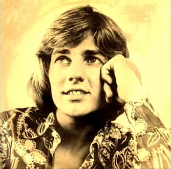 Imagen sepia de un joven Sergio Denis con cabello castaño claro, camisa estampada con cuello, y la mano izquierda apoyada cerca de la mejilla