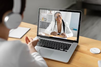 Una persona con auriculares frente a un portátil. En la pantalla, una médica afroamericana sonríe con gafas y bata blanca, ofreciendo una consulta virtual