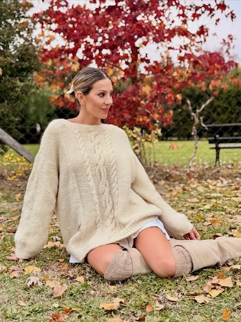 Noelia Marzol sentada en la hierba con hojas secas, viste un suéter de punto color crema y botas beige, con un árbol de hojas rojas al fondo