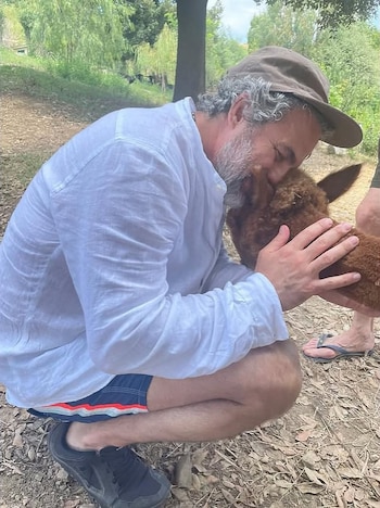 Mark Ruffalo, agachado al aire libre con camisa blanca y gorra, sostiene la cabeza de una pequeña alpaca marrón