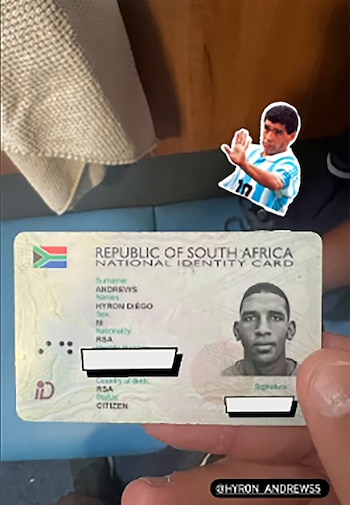 Una mano sostiene un documento de identidad de Sudáfrica a nombre de Hyron Diego Andrews junto a una figura recortada de Diego Maradona