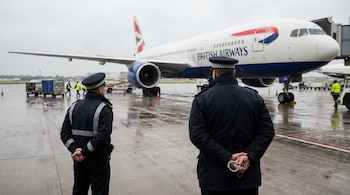 Dos oficiales de policía, vistos de espaldas, con uniformes oscuros en una pista mojada de aeropuerto; un avión de British Airways está al fondo.