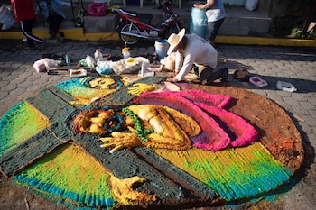 Un hombre con gorra azul crea una alfombra de aserrín colorida en el suelo, con diseños de un sol radiante y dos estructuras arquitectónicas coloniales, en una calle empedrada