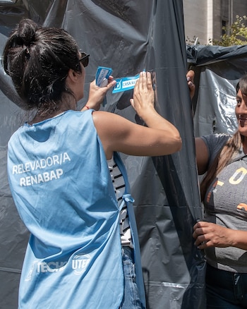 Una mujer con chaleco azul y gafas de sol, con el pelo recogido, fija una pegatina azul de 'Casa Registrada' en una lona negra; otra mujer sonríe a su derecha