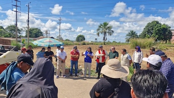 Grupo de personas reunidas en un campo abierto. Una mujer con chaleco azul habla con micrófono, rodeada de hombres. Al fondo, postes, árboles y una sombrilla publicitaria