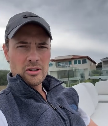 Un hombre con gorra deportiva y chaleco polar gris mira hacia el frente desde una terraza con muebles blancos y viviendas al fondo