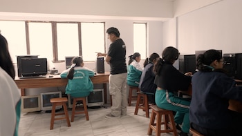 Un hombre señala una pantalla de computadora mientras estudiantes usan PC en un aula. Hay ventanas, escritorios de madera y monitores antiguos en el suelo