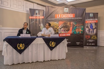 Dos hombres en una mesa con mantel azul y dorado, frente a banners de vinilo con información de CONAP sobre la protección de la fauna silvestre