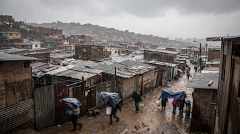 Asentamiento humano en ladera con casas de madera y techos de chapa, personas caminan con lonas y baldes en calle de tierra y barro bajo lluvia intensa