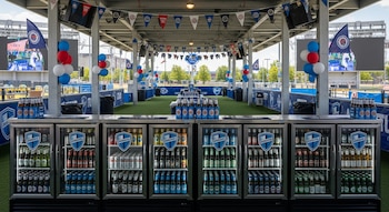 Un espacio abierto decorado con banderas, globos y pantallas, donde varias barras exhiben refrigeradores repletos de botellas y latas de cerveza.