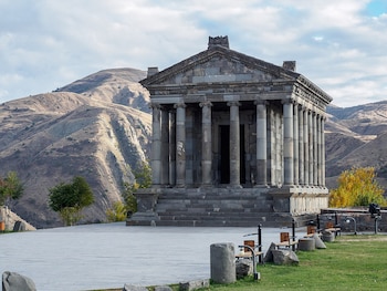 Templo de piedra con columnas clásicas y escalinata, situado en una plaza adoquinada con césped y colinas de fondo bajo un cielo despejado