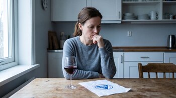 Una mujer sentada en una mesa de cocina de madera, mirando hacia abajo. Frente a ella hay una copa de vino tinto y un papel con el logo azul marino de Alcohólicos Anónimos.