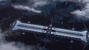 Vista aérea de una gran estructura blanca y gris de forma simétrica sobre una masa de agua, rodeada por áreas de tierra oscura y cubierta por nubes