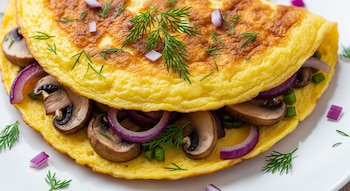 Primer plano de un omelette plegado con champiñones, aros de cebolla morada y eneldo fresco esparcido sobre él y alrededor en un plato blanco.