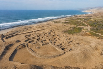 Vista aérea de estructuras de piedra antiguas en un terreno desértico junto al mar y una playa de arena.
