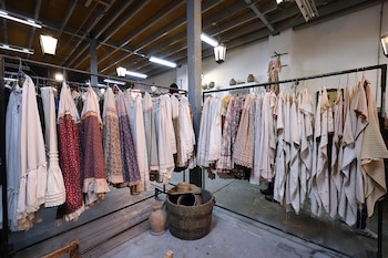 Percheros metálicos cargados con vestidos de tela clara y estampada, junto a un barril de madera y vasijas en un almacén