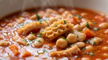 Guiso de mondongo, porotos, garbanzos, trozos de zanahoria, perejil picado, caldo rojo, gotas de aceite, vapor saliendo de un plato.