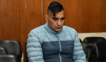 Hombre con cabello oscuro y chaqueta acolchada azul mira hacia abajo, sentado frente a una pared de madera y sillas negras