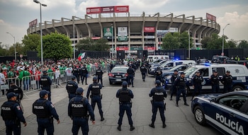 Estadio Azteca, agentes de policía uniformados, vehículos policiales, vallas metálicas, gran multitud de personas, árboles, cielo nublado.