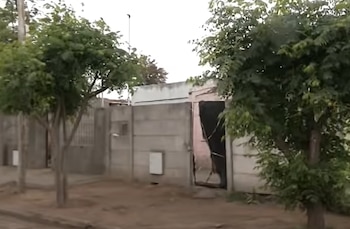 Un muro de hormigón con una entrada abierta, medidores de servicios públicos y árboles en la acera frente a una casa