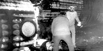 Dos personas, una agachada y otra de pie, dentro de la estructura metálica dañada de un avión. Hay restos esparcidos en el suelo