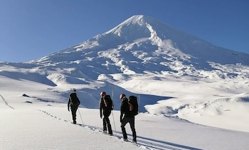 Vista trasera de tres personas con mochilas y bastones de trekking, caminando sobre un terreno cubierto de nieve con huellas, hacia una gran montaña nevada bajo un cielo azul brillante
