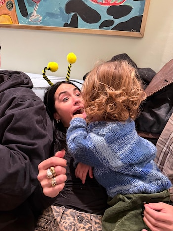 Una mujer con diadema de abeja y un niño pequeño de cabello rizado se miran. El niño toca la cara de la mujer con la lengua. Se aprecian anillos en los dedos de la mujer, un sofá y una pintura abstracta al fondo