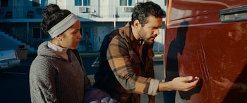 Primer plano de una mujer (Lou Llobell) y un hombre de perfil, mirando las profundas marcas de arañazos en la pintura roja de un vehículo, con un motel de fondo