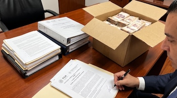 Un hombre firma un documento en un escritorio de madera junto a una caja de cartón llena de fajos de billetes de doscientos pesos y varios archivadores.