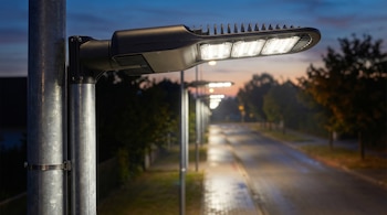 Primer plano de un moderno farol LED negro sobre poste metálico. Una hilera de faroles LED idénticos ilumina una calle mojada al anochecer, bajo un cielo azul oscuro.