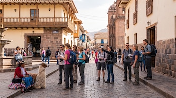 Calle adoquinada con edificios coloniales, una iglesia de piedra, una fuente. Un grupo de turistas con mochilas y una mujer local sentada junto a una llama